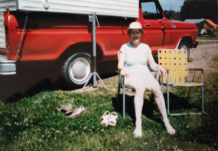 a vintage photo of a grandmother sitting on a lawn chair with her bare feet in the grass. Behind her is a truck camper hoisted on back of a red Ford truck.