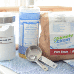 A glass jar of washing soda sits next to a bottle of Dr Bronner's Castile Soap and Simply Clean Borax. In the foreground sits stainless steel measuring cup and spoon.
