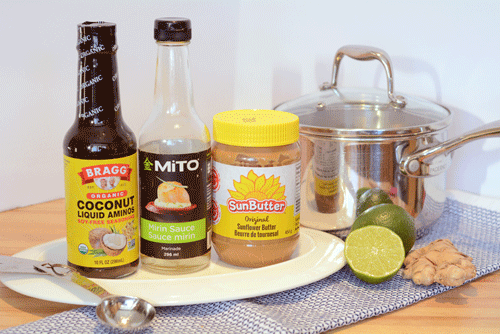 Gluten free, soy free, dairy free products are the main subjects of the photo. Coconut aminos, mirin and SunButter bottles sit on a white serving platter. In the background is a saucepan, a half of a lime and a piece of ginger. 