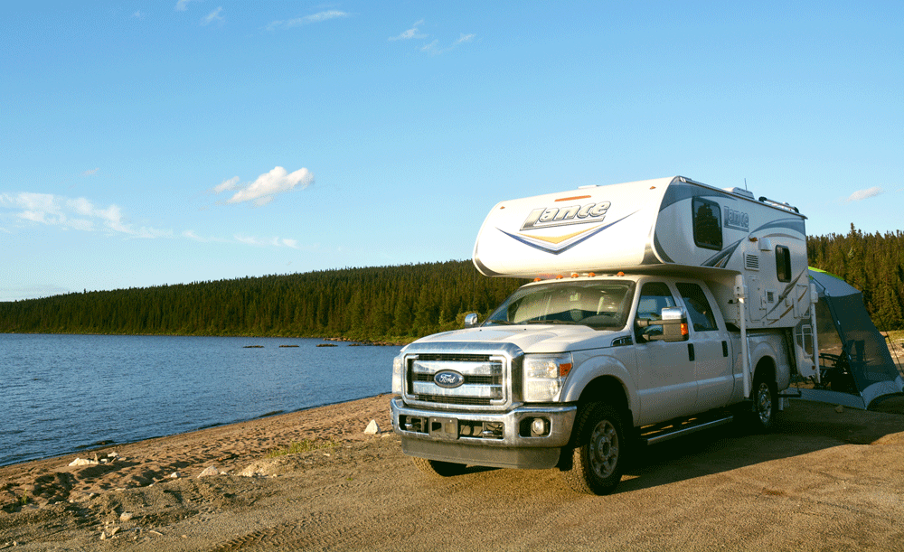 Photo of a Lance 825 truck camper sitting on top of a white Ford F-150 truck on a beach. There is water and trees in the background and a clear blue sky.