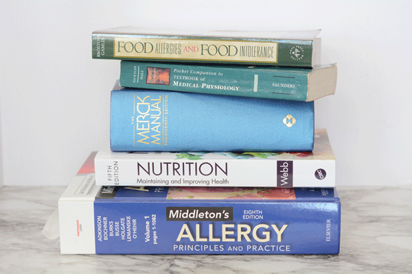 A pile of medical text books are stacked on a marble countertop. Among the titles are "Middleton's Allergy Principles and Practice", "Nutrition: Maintaining and Improving Health," the "Merck Manual", "Pocket Companion to Textbook of Medical Physiology," and "Food Allergies and Food Intolerance".