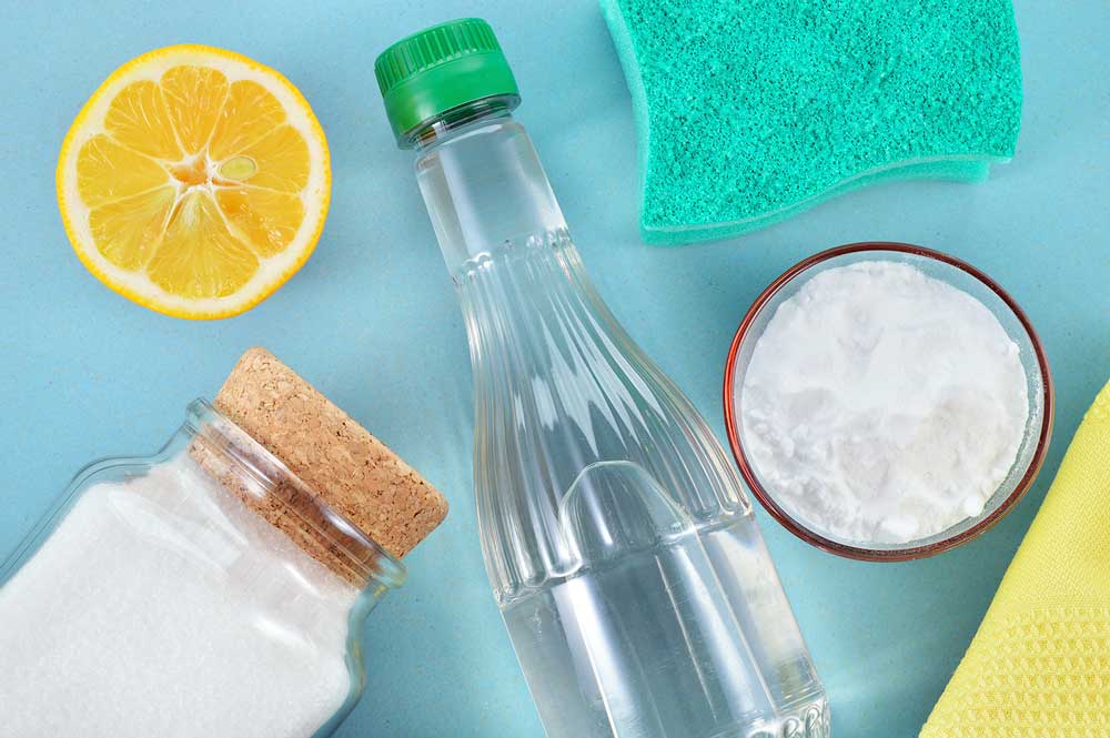 An overhead shot of assorted household cleaning ingredients laid on a light blue background. On top sits a half a lemon, a clear bottle of vinegar and a green sponge. Underneath is a clear glass bottle of salt with a cork and a dish of baking soda and a yellow cleaning cloth.
