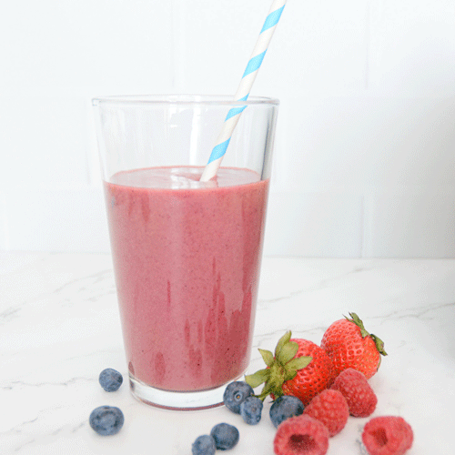 A large pink smoothie in a tall clear glass with a striped white and blue paper straw sits on a white marble countertop. Fresh strawberries, raspberries and blueberries surround the glass.