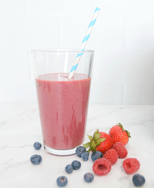 A large pink smoothie in a tall clear glass with a striped white and blue paper straw sits on a white marble countertop. Fresh strawberries, raspberries and blueberries surround the glass.