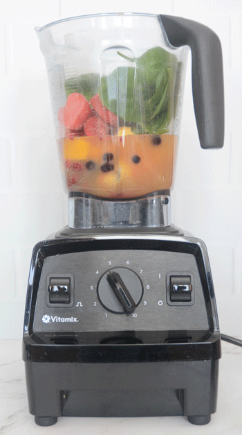 A Vitamix blender full of fruit juice, frozen berries and fresh spinach sitting on a white marble countertop.