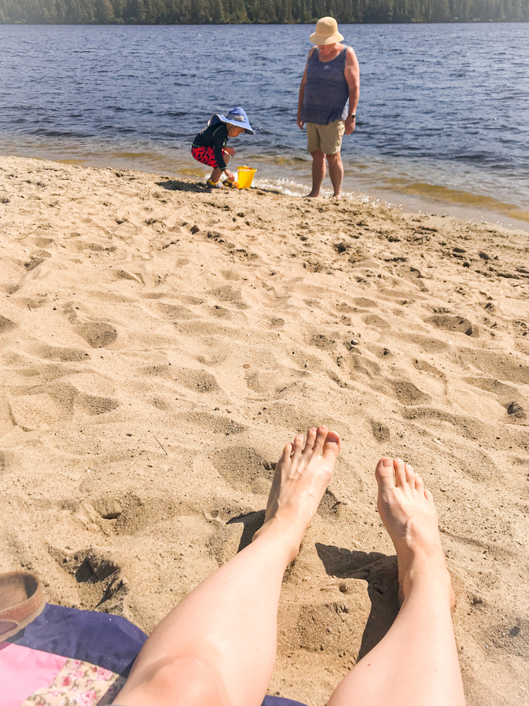 A photo of a sandy beach. The feet of one person is visible at the bottom of the frame. A small child and senior lady are playing by the water’s edge.