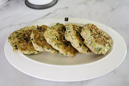 A row of five fried Allergen Friendly Chicken Burger patties are displayed horizontally on a white oval plate atop a white marble countertop. 