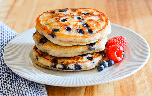 Three gluten-free vegan blueberry pancakes are stacked on a white and grey plate. To the right of the pancakes sit two raspberries and a few fresh blueberries. The plate sits on a wooden countertop. A blue and white patterned cloth is visible in the bottom left corner.
