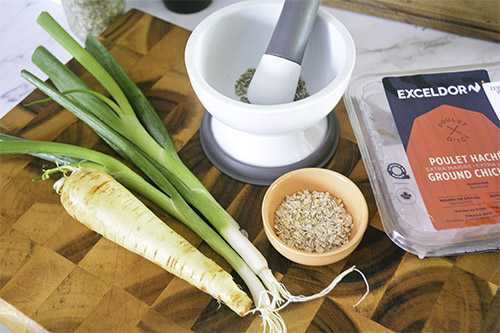 Ingredients for Allergen-Friendly Chicken Burger patties are laid out on a wooden cutting board. In the centre is a white and grey mortar and pestle containing fennel seeds. To the right is a plastic package of ground chicken. To the far left sits a beige parsnip, two stalks of green onion and a small peach coloured bowl of quick oats.
