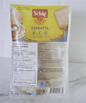 A package of four Schar gluten free, vegan Ciabatta buns sit in sealed plastic packaging atop a white marble countertop.