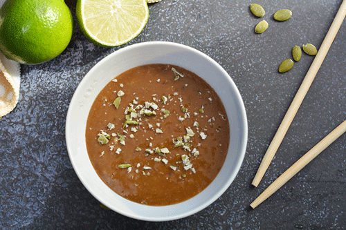 A small bowl of Allergen-Free Thai Faux-Nut Sauce sits on a dark grey countertop. There are bright green limes in the top left corner of the image. One of them is cut in half. Two wooden chopsticks are placed to the right of the bowl. Green pumpkin seeds are scattered on the counter and crushed seeds are sprinkled on the sauce.