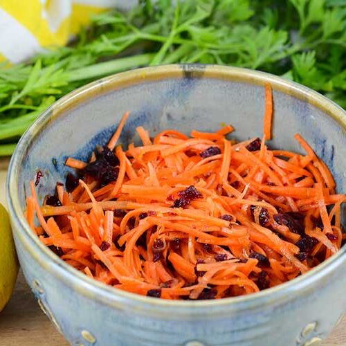 A light blue artisan bowl sits on top of a wooden cutting board. The bowl is filled with Cranberry Carrot Slaw. To the left of the bowl sits a bright yellow lemon. In the background, green carrot fronds sit next to a yellow and white striped cup towel.