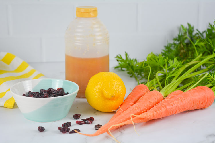 Ingredients for the Cranberry Carrot Slaw recipe sits on a white marble countertop. Among these are four freshly picked carrots, a lemon, a container of honey and a small white and turquoise bowl of dried cranberries. There is a striped yellow and white cup towel in the background and some dried cranberries sprinkled in the foreground of the image. 