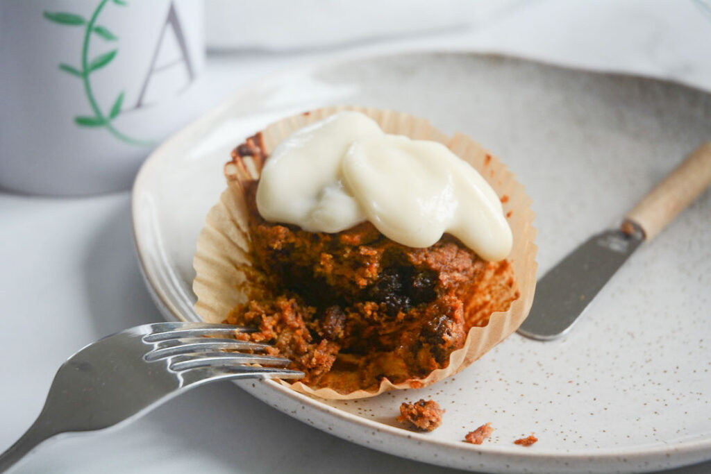 A gluten free vegan carrot muffin sits partially eaten on a light grey artisanal plate. There is a dollop of white dairy free cream cheese atop the muffin. A small spreading knife and a fork sit on the plate amid crumbs. There sits a white mug in the upper left corner of the frame with the green and grey Allergen Free Artistry logo.
