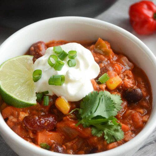 A close-up shot of Epic Vegan Chili in a white bowl sitting on a light grey wooden tabletop. The chili is topped with a dollop of vegan sour cream, a sprinkling of chopped green onion, a garnish of fresh cilantro sprigs and a lime wedge. There is a red hot pepper in the upper left of the shot and a spoon on the left of the bowl.