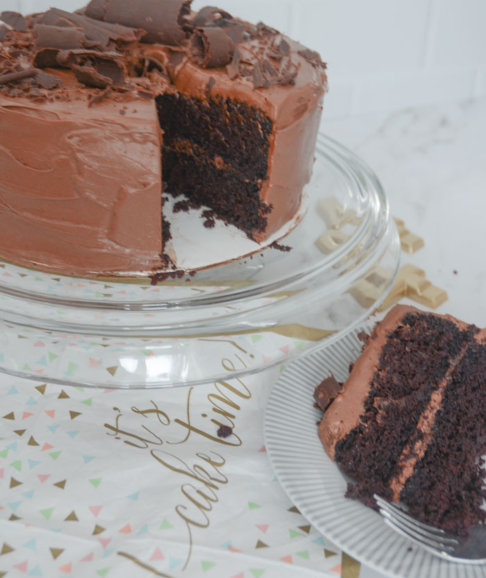 A gluten free dairy free chocolate cake frosted with dairy free chocolate "cream cheese" icing and topped with chocolate shavings sits on a a glass cake stand. One piece is cut out and placed on a grey and white striped plate in the foreground. On the table is a paper napkin that reads "it's cake time" in gold cursive. There are pastel coloured triangle confetti in the graphics. 