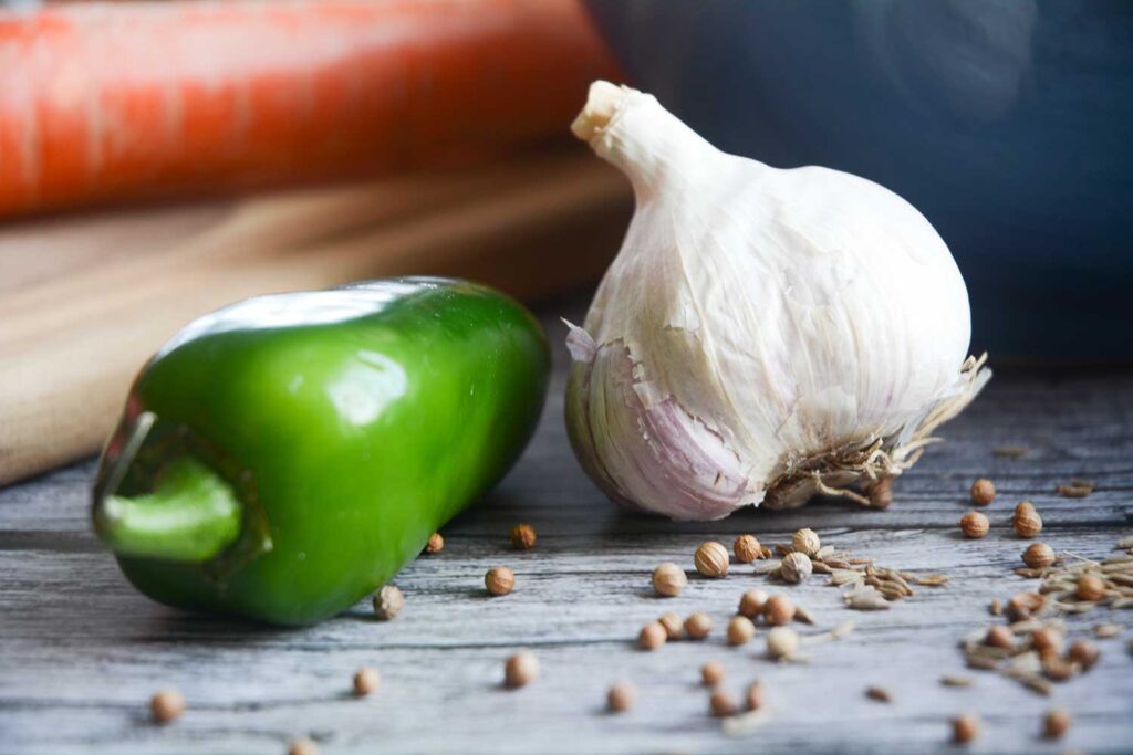 A close up shot of a green jalapeno pepper and a bulb of garlic. Sprinkled in the foreground of the shot are coriander and cumin seeds. 
