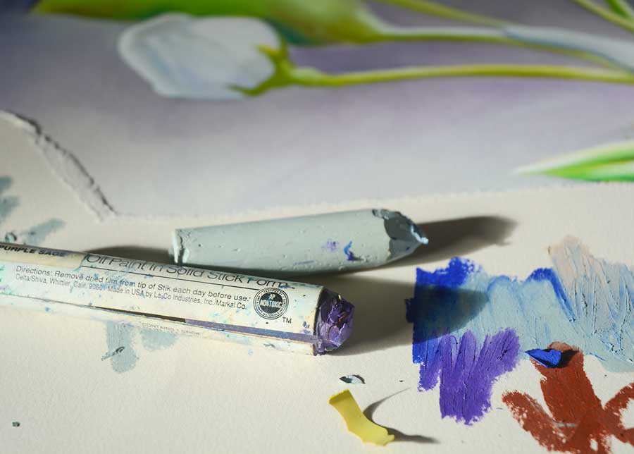 Two oil sticks are placed on a sheet of rag paper. The one in the foreground is purple with a white wrapper. The one behind is a light blue colour. There is experimental blending of oil colours on the paper. A  blurred painting of a flower is visible in the background of the image.