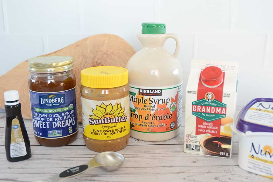 An assortment of allergy friendly pantry items sit on a wooden counter. Among them is a small plastic bottle of "Great Value" vanilla extract, "Lundberg Sweet Dreams" brown rice syrup in a glass bottle, "SunButter" Sunflower Butter in a plastic jar with a yellow lid, "Kirkland" maple syrup in a plastic jug, "Grandma" molasses in a small carton and "Nuvel" vegan margarine in a white and purple plastic tub. 