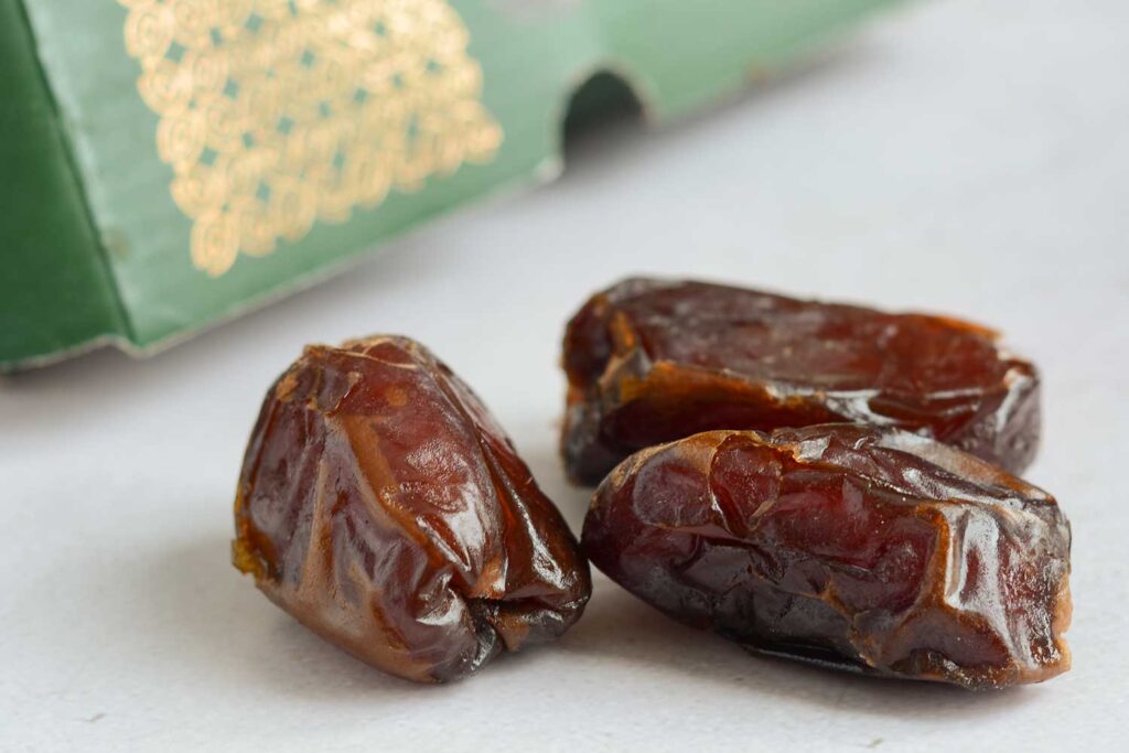 A close up shot of three brown Medjool dates sitting on a white countertop. Out of focus in the background is a green box with gold patterning.