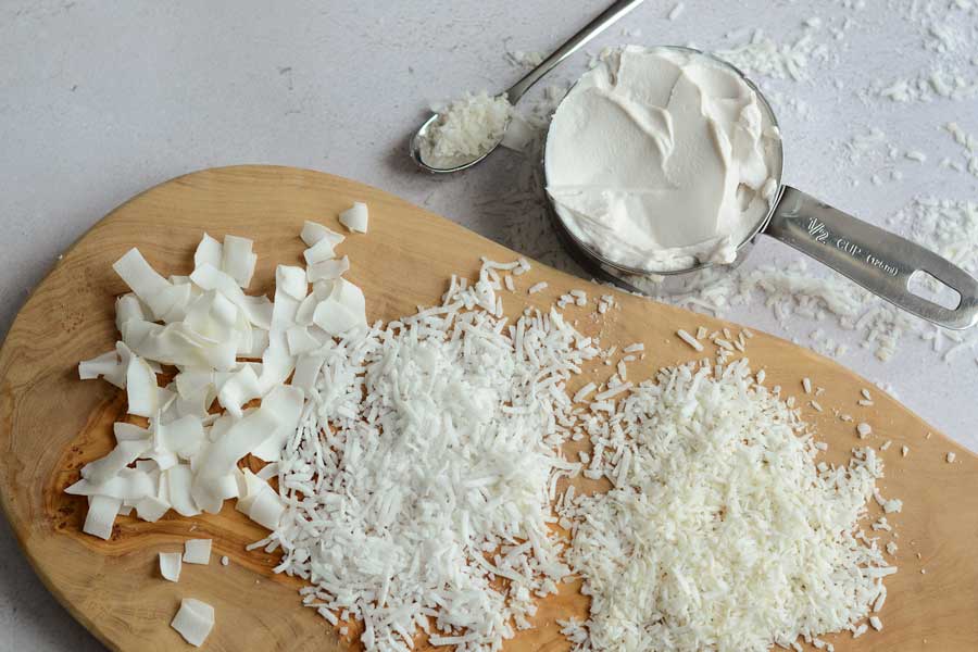 A free-formed golden brown wooden cutting board sits on a white countertop. On the board in three separate piles are a variety of coconut. On the far left sits coconut flakes, the middle is long shredded sweetened and the far right is finely grated. On the top of the photo sits a stainless steel 1/2 cup measure holding coconut cream. To the left of it is a mini spoon holding shredded coconut. There is shredded coconut scattered on the counter.