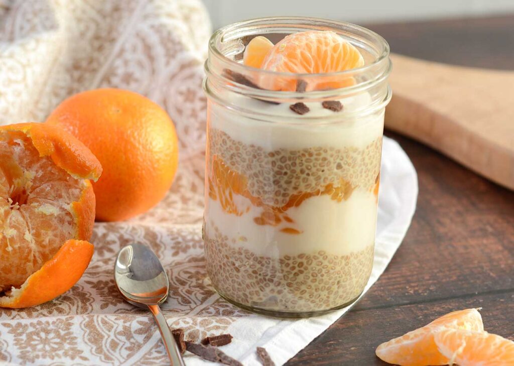 A 250 ml glass jar containing Orange Creamsicle Chia Pudding sits on a decorative white and gold patterned fabric atop a dark wooden counter top. In the jar is layered white chia pudding, coconut yoghurt and clementines, topped with two clementine segments and some dairy-free chocolate shavings. to the left of the jar sits a small stainless steel spoon and two clementines. One of the fruits is partially peeled.