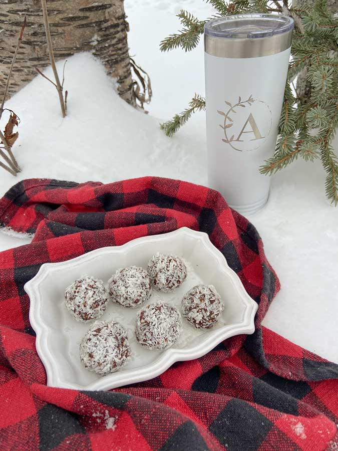 Six vegan chocolate coconut snowball cookies sit on a decorative white scalloped dish. This is placed on a black and white plaid fabric, on the snow. There is a tall white tea caraffe with the Allergen Free Artistry logo on it. A tree trunk and spruce sprigs are visible in the background. 