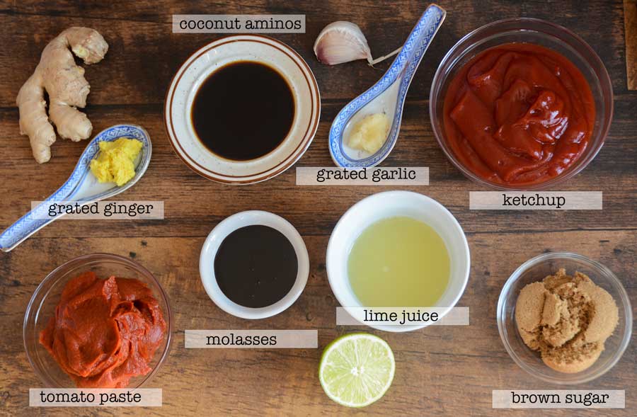 An overhead shot of labelled ingredients for gluten free and dairy free Best Asian BBQ Sauce Recipe. Included in the image is grated ginger, coconut aminos, grated garlic, ketchup, tomato paste, molasses, lime juice and brown sugar in various coloured bowls and spoons. All sit atop a wooden countertop