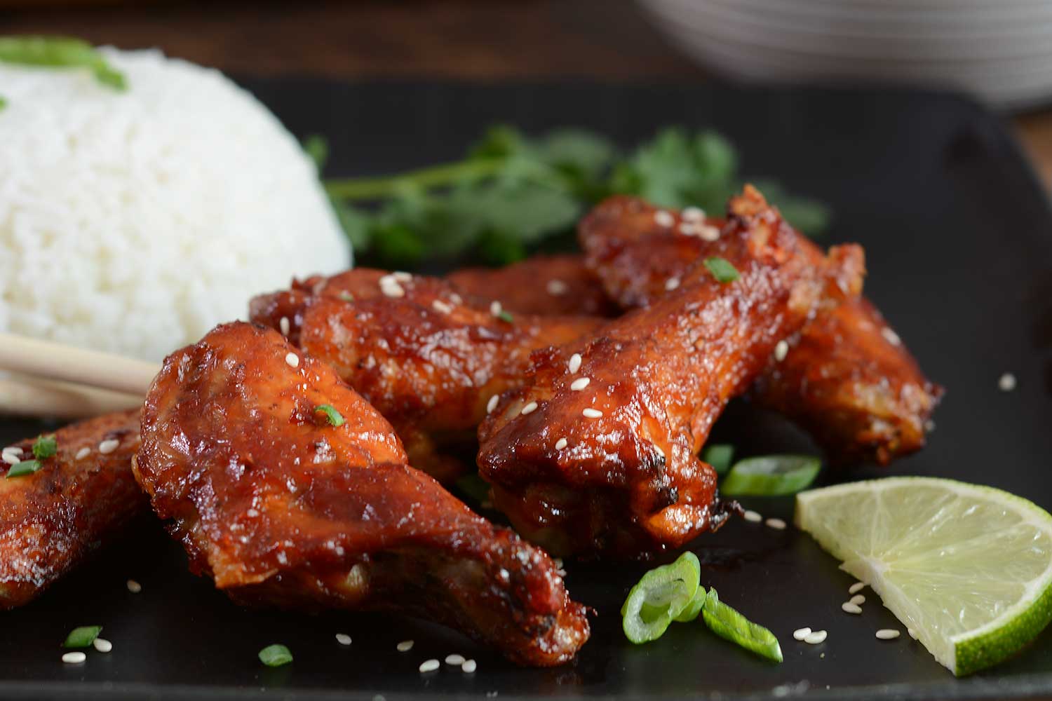 A close up of Asian Bbq Chicken Wings piled on a black serving plate. A mound of jasmine rice and sprig of cilantro is out of focus in the background. There is a half slice of lime in the foreground. Fresh spring onion and sesame seeds are sprinkled on the dish.