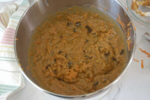 A process shot of the Top 8 Allergen-Free Carrot Cake batter in a stainless steel mixing bowl. There is a cream, pink, yellow and green striped hand towel on the left of the bowl.
