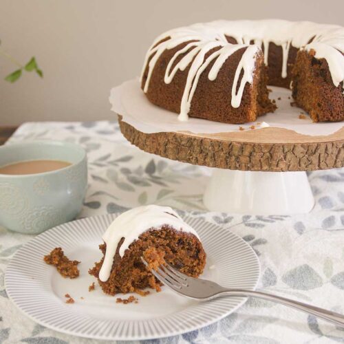 A Top 8 Allergen-Free Carrot Cake drizzled with Dairy Free "Cream Cheese" Glaze is placed on a rustic wooden cake stand. In the foreground sits a piece of carrot cake on a grey and white patterned plate. There is a steel fork placed on the plate holding cake crumbs. There is a turquoise mug holding tea to the left of the shot. All is placed on top of a blue, green and white patterned table cloth.