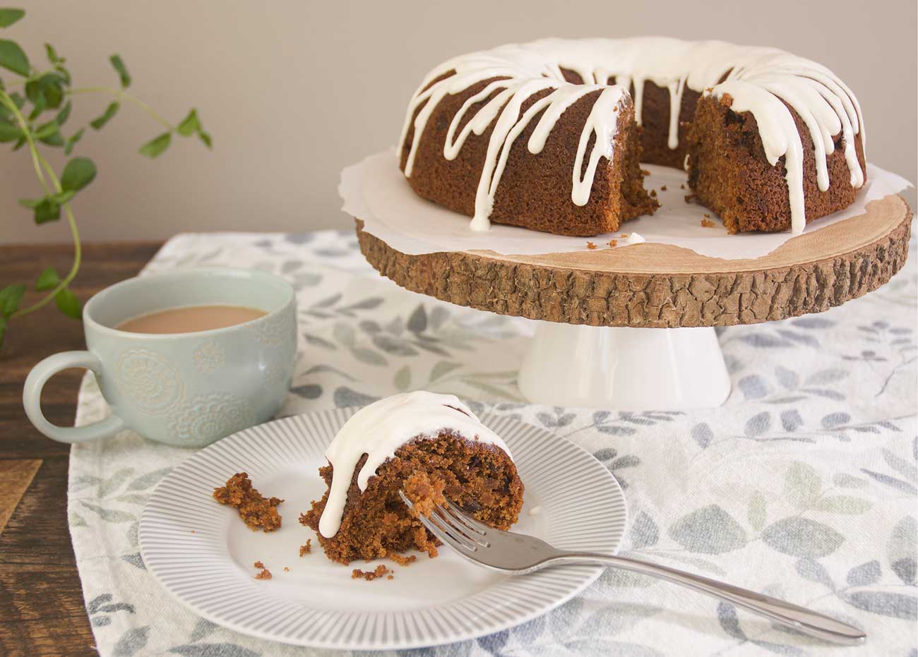 A Top 8 Allergen-Free Carrot Cake drizzled with Dairy Free "Cream Cheese" Glaze is placed on a rustic wooden cake stand. In the foreground sits a piece of carrot cake on a grey and white patterned plate. There is a steel fork placed on the plate holding cake crumbs. There is a turquoise mug holding tea to the left of the shot. All is placed on top of a blue, green and white patterned table cloth.