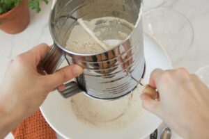 A process shot showing the sifting of dry ingredients for the Top 8 Allergen-Free Carrot Cake recipe. A pair of hands hold the flour sifter over a white ceramic bowl.