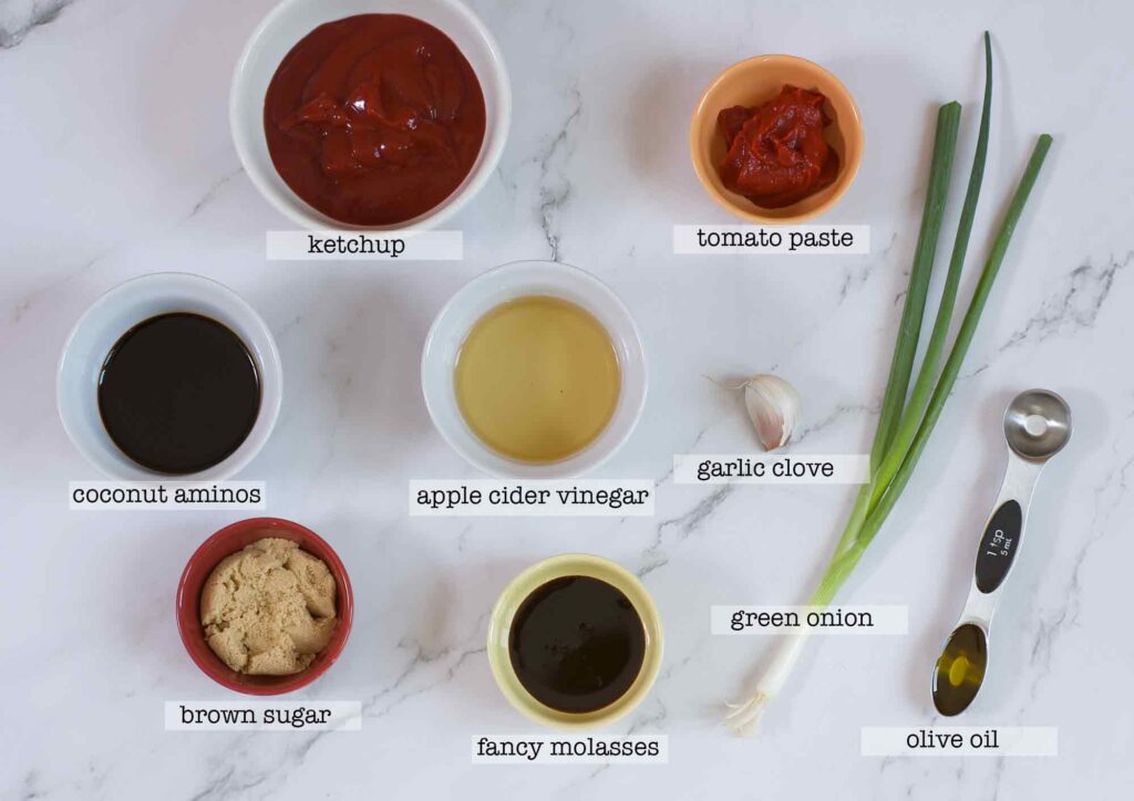 An overhead shot of ingredients for the Sweet and Tangy Allergen Free BBQ Sauce Recipe. Bowls are arranged on top of a white marble surface. There is text beneath each, naming the ingredient. On the top is ketchup and to the right, tomato paste. The middle row from left to right is coconut aminos, apple cider vinegar and a garlic clove. The bottom row has brown sugar, fancy molasses a green onion and a measuring spoon holding a teaspoon of olive oil.