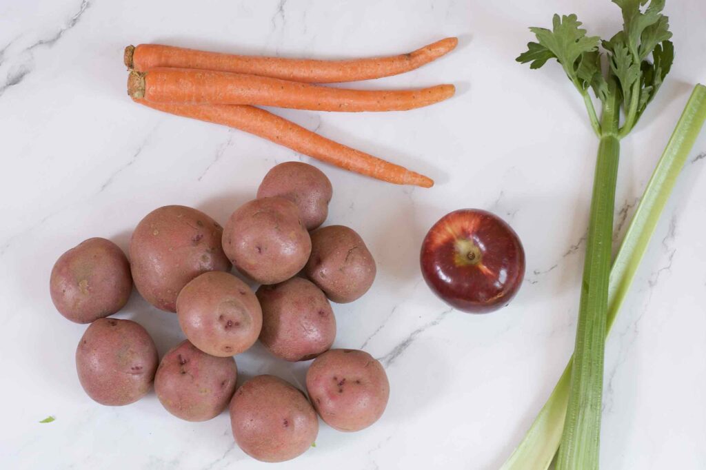 Fresh produce laid out on a white marble countertop. Three unpeeled carrots lie at the top of the image, a pile of red skinned potatoes sit underneath. In the center, a red gala apple and to the right, two stalks of celery.