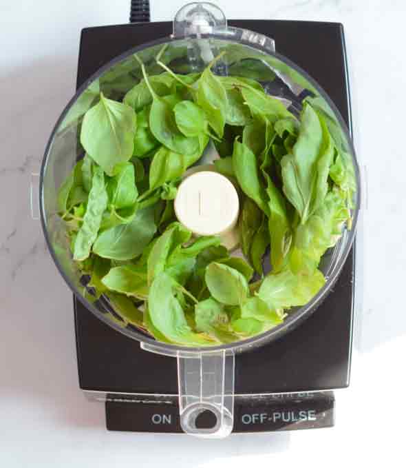 An overhead view of fresh basil and baby spinach leaves in a cuisinart food processor sitting on a white countertop.