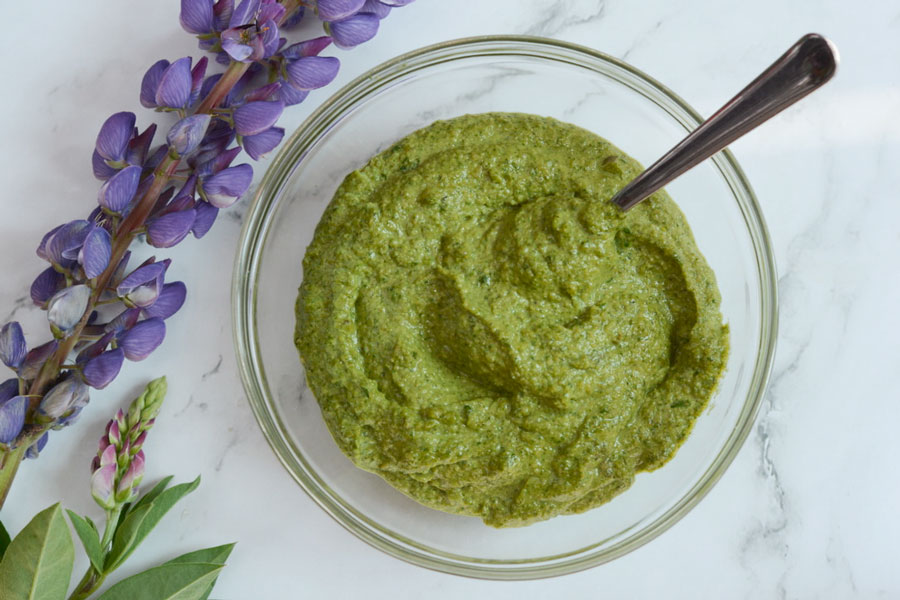 An overhead shot of vibrant green Perfect Pesto in a clear glass bowl with a spoon sticking out. It is sitting on a white marble countertop. To the left of the bowl is a contrasting purple lupin flower.